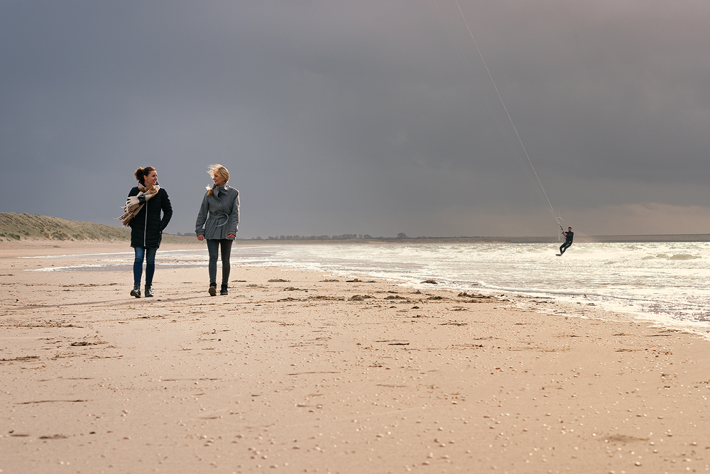 Twee mensen wandelen op winderig strand met kitesurfer